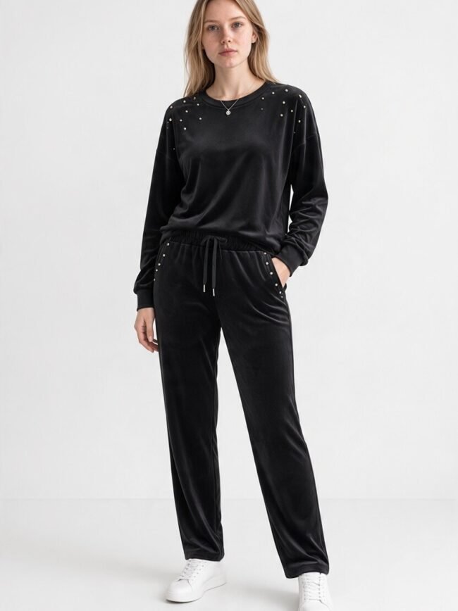 Full-body shot of a model in a black velour tracksuit with stud embellishments and white sneakers, standing with hands in pockets.