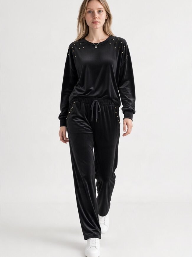 Front view of a woman wearing a black velour tracksuit with gold studs on the shoulders and pockets, paired with white sneakers.