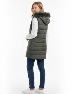 Woman wearing a knee-length olive green puffer vest with a fur-trimmed hood, white long-sleeve top, blue jeans, and white sneakers, facing slightly to the side.