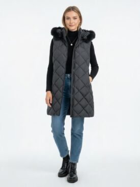 Woman wearing a dark quilted vest with a faux-fur hood over a black turtleneck, blue jeans, and black lace-up boots, standing with hands in pockets in a studio setting.