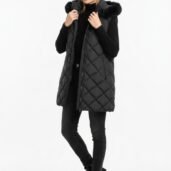 Woman wearing a long black quilted puffer vest with a faux-fur hood over a black turtleneck, black jeans, and black ankle boots, posing against a white background.
