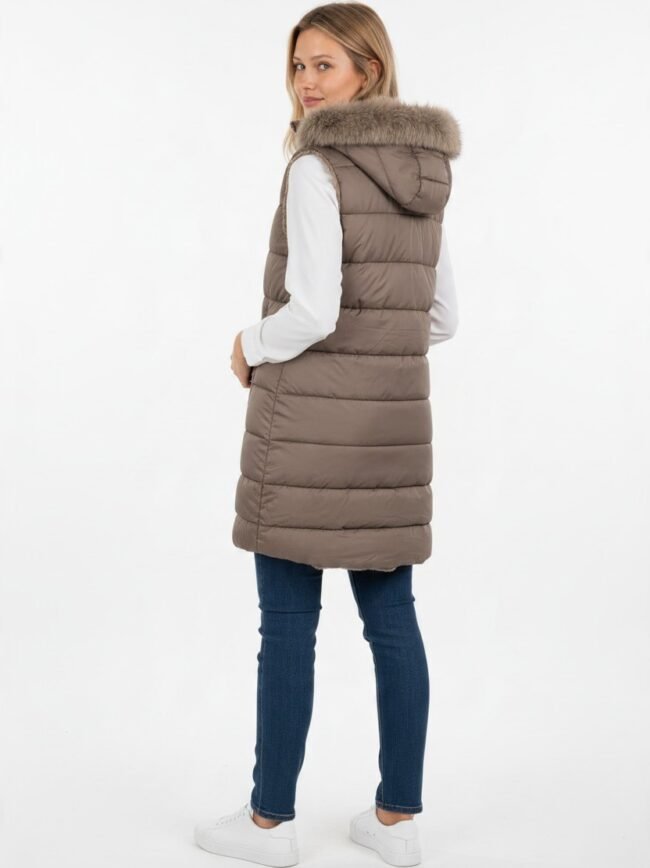 Side-view of a woman wearing a taupe quilted puffer vest with a fur-trimmed hood over a white long-sleeved top, blue jeans, and white sneakers.