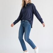 Young woman in a navy button-down shirt, blue jeans, and white sneakers, posing mid-step in a studio.