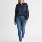 Woman wearing a navy button-down blouse with blue jeans and white sneakers, standing in a bright studio setting.