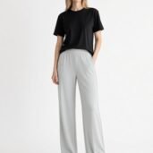 Woman wearing a black t-shirt and light gray wide-leg pants, standing with one hand in her pocket against a white background.