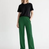 Full-body shot of a woman in a black T-shirt, emerald green wide-leg pants, and white sneakers against a light backdrop, relaxed pose with hands in pockets.