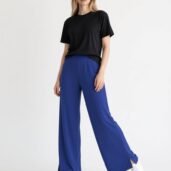 Model wearing a black T-shirt and royal blue wide-leg pants with white sneakers, standing in a studio on a white background.