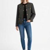 Woman wearing a black-and-cream tweed-style jacket with gold buttons, black top, blue jeans, and white sneakers.