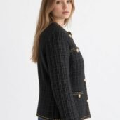 Profile of a woman wearing a black textured tweed jacket with gold trim and buttons, against a light background.