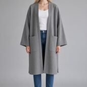 Woman standing in an oversized gray coat over a white top and blue jeans with white sneakers against a neutral backdrop