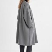 Woman in a loose gray coat standing in profile against a light background, looking at the camera.