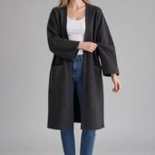 Woman standing in a dark long coat over a white top and blue jeans, wearing white sneakers.