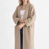 Woman wearing a beige long coat with large front pockets over a white top and blue jeans, standing against a plain white background.