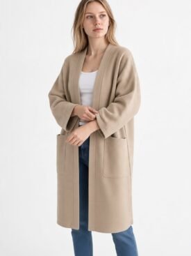 Woman wearing a beige long coat with large front pockets over a white top and blue jeans, standing against a plain white background.