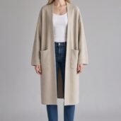 Woman standing in a light beige, open-front coat with two large pockets, white top, blue jeans, and white sneakers.