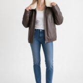 Woman wearing a brown shearling‑lined leather jacket, white top, blue jeans, and white sneakers against a light background.