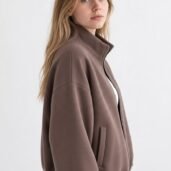 Side profile of a young woman wearing a brown zip-up jacket against a pale background.