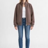 Woman wearing a taupe-brown bomber jacket, white top, blue jeans, and white sneakers, standing against a white background.