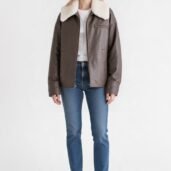 Model wearing a brown shearling-collar leather jacket over a white tee and blue jeans, white sneakers, against a white background.