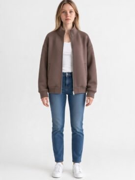 Woman wearing a taupe-brown bomber jacket, white top, blue jeans, and white sneakers, standing against a white background.