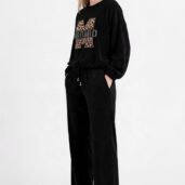 Woman wearing a black velour tracksuit with leopard-print M logo on the sweatshirt and white sneakers, standing with hands in pockets.