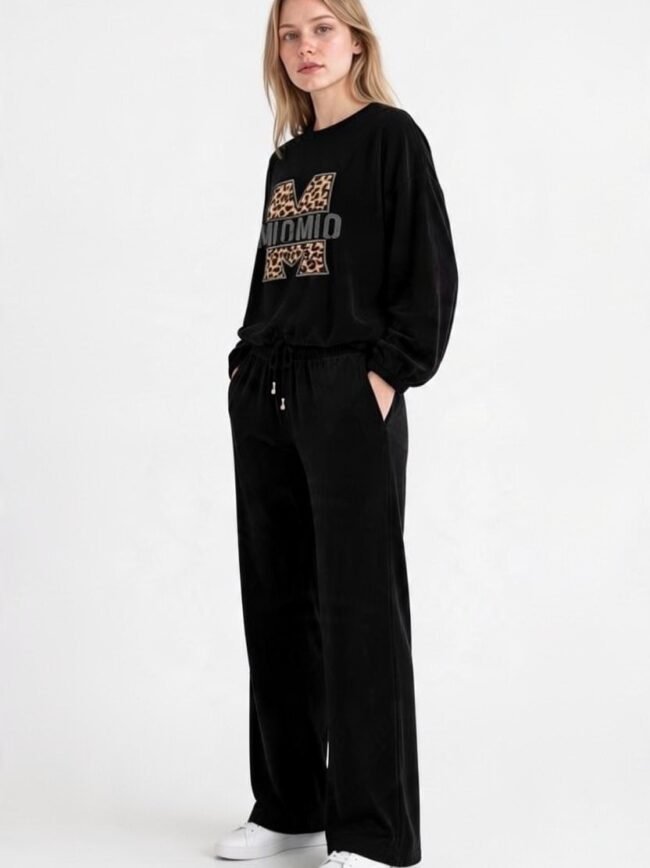 Μαύρο Σετ 3 2 Woman wearing a black velour tracksuit with leopard-print M logo on the sweatshirt and white sneakers, standing with hands in pockets.
