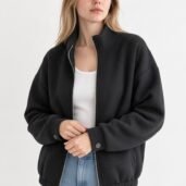 Woman wearing a black zip-front bomber jacket over a white top, with light-wash jeans, standing against a pale background.