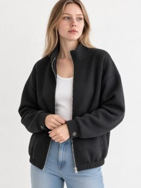 Woman wearing a black zip-front bomber jacket over a white top, with light-wash jeans, standing against a pale background.