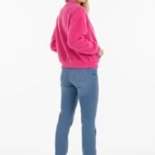 Woman standing in a pink sweater and blue jeans, turned slightly to the side, facing camera with a subtle smile, white sneakers