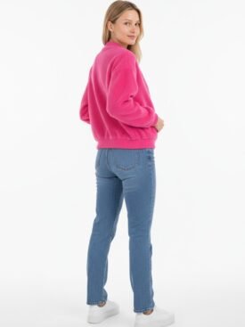 Woman standing in a pink sweater and blue jeans, turned slightly to the side, facing camera with a subtle smile, white sneakers