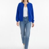 Full-body portrait of a woman in a royal blue bomber jacket, white top, light-wash jeans, and white sneakers against a white studio background, walking toward the camera.