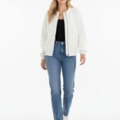 Full-body portrait of a woman in a casual outfit: white zip-front jacket, black top, blue jeans, and white sneakers in a bright studio setting.