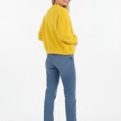Woman in a bright yellow knit sweater, blue cuffed jeans, and white sneakers poses with her back to the camera, turning slightly to smile over her shoulder on a white backdrop.