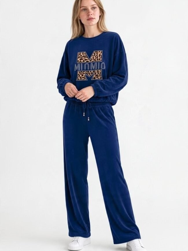 Μπλε Σκούρο 1 Full-body shot of a woman in a blue velour tracksuit with a leopard-print M logo and white sneakers, standing casually.