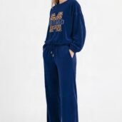 Woman in a blue velour tracksuit with a leopard-print 'M' graphic on the top and white sneakers, standing with hands in pockets.