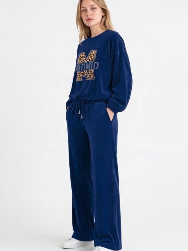Μπλε Σκούρο 2 Woman in a blue velour tracksuit with a leopard-print 'M' graphic on the top and white sneakers, standing with hands in pockets.