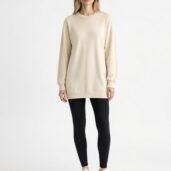 Woman modeling a beige oversized sweater dress with black leggings and white sneakers, standing against a plain white background.