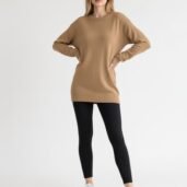 Woman wearing an oversized camel sweater tunic, black leggings, and white sneakers, standing with hands on hips.