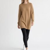 Woman models a camel-colored knit sweater dress over black leggings and white sneakers in a white studio setting.