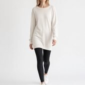 Woman walking toward the camera in an oversized cream knit sweater, black leggings, and white sneakers in a white studio setting.