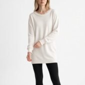 Woman wearing an oversized cream knit sweater with scalloped cuffs, paired with black tights, standing against a white background.