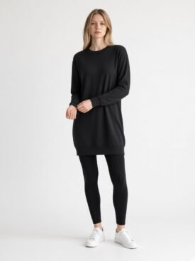 Woman wearing a black long-sleeve tunic sweater, black leggings, and white sneakers against a light background, posing casually.