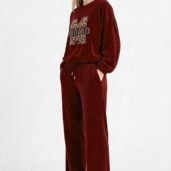 Woman in burgundy velour loungewear set with a drawstring waist, hands in pockets, wearing white sneakers against a plain light background.