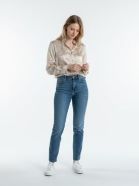 Full-body shot of a woman in a beige patterned blouse, blue high-waisted jeans, and white sneakers, standing against a white studio backdrop while adjusting her sleeves.