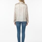 Woman seen from behind wearing a light beige patterned blouse, blue jeans, and white sneakers, standing against a plain white backdrop