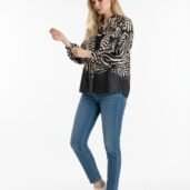 Woman in a zebra-print blouse, blue jeans, and white sneakers adjusting her sleeve against a white backdrop.
