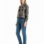Young woman standing against a white background, wearing a black-and-beige animal print blouse, blue jeans, and white sneakers.