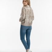 Woman standing with her back turned slightly toward the camera, wearing a beige patterned jacket, blue jeans, and white sneakers against a plain white background.