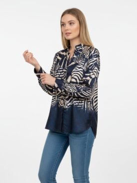 Woman in a navy beige animal-print blouse and blue jeans, standing against a white background, adjusting her sleeve.