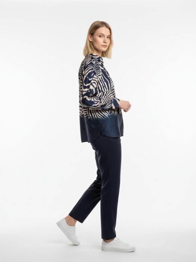 Side view of a woman wearing a navy and white zebra-print blouse, navy trousers, and white sneakers in a studio setting.